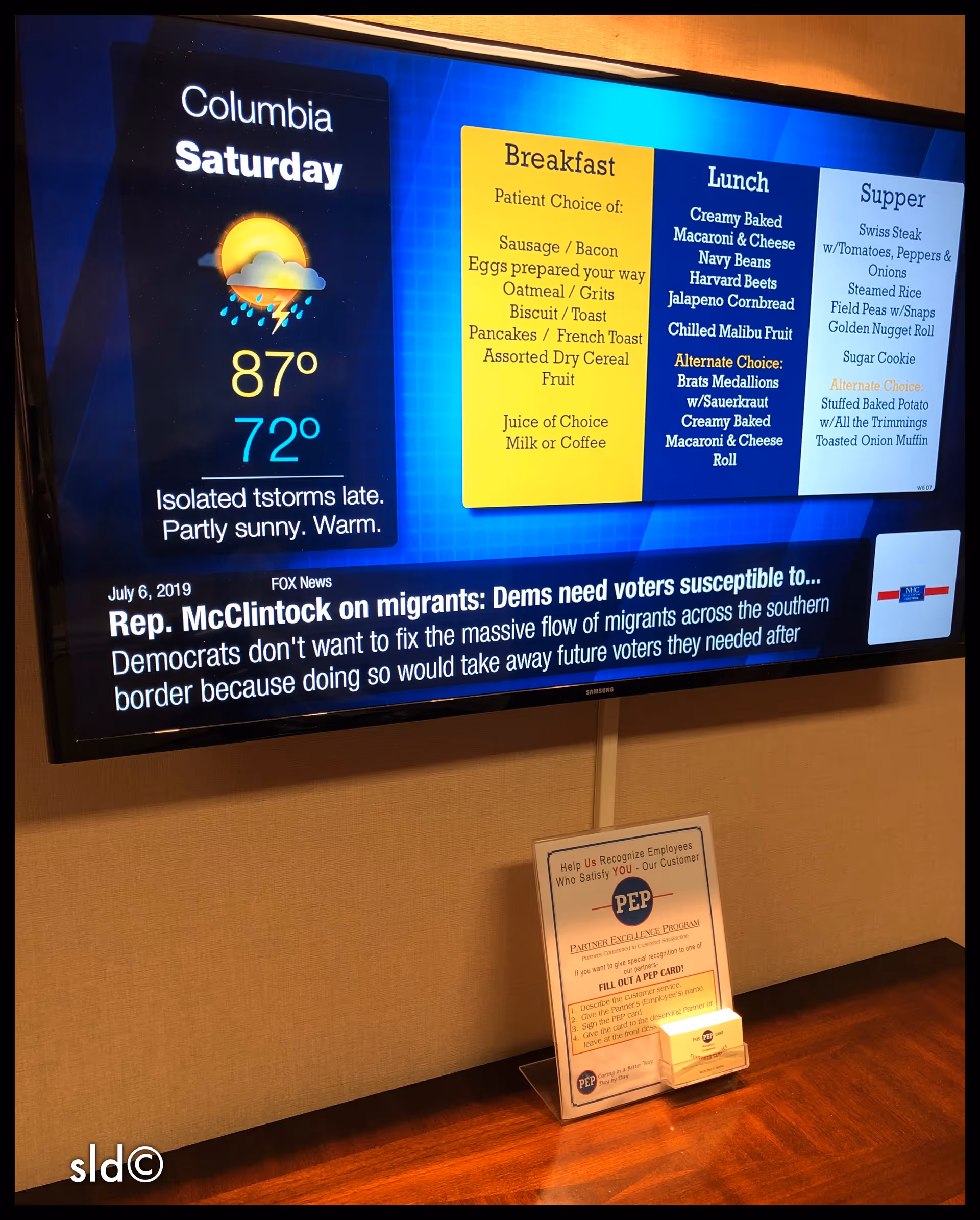 Wall-mounted TV displays weather, breakfast and lunch menus, with a small informational sign nearby.