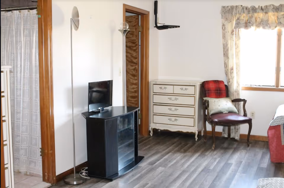 Bedroom in senior living facility with dresser, TV stand, chair, and window blinds.