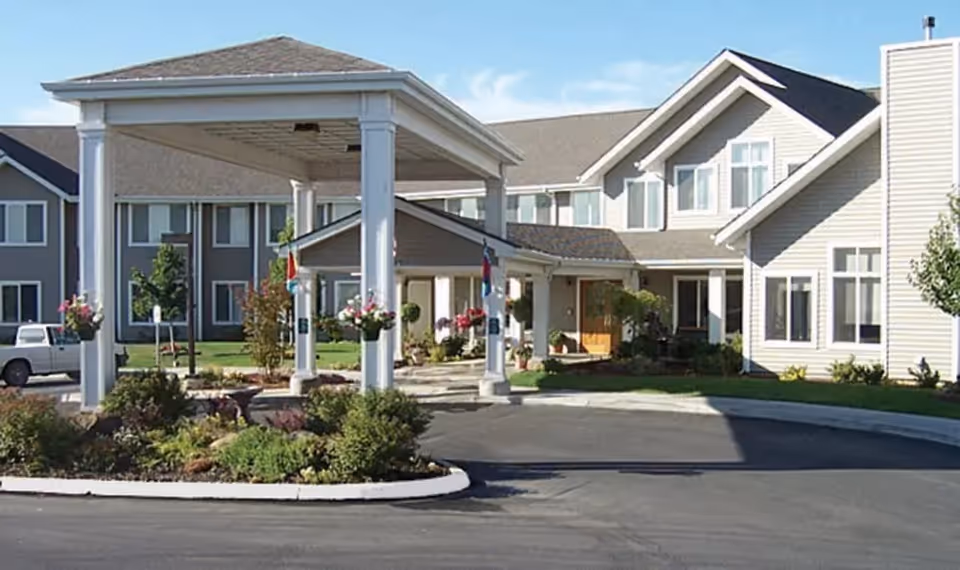 Front entrance of Bishop Place Senior Living with a covered porte-cochere, landscaped circular drive, and hanging flower baskets.