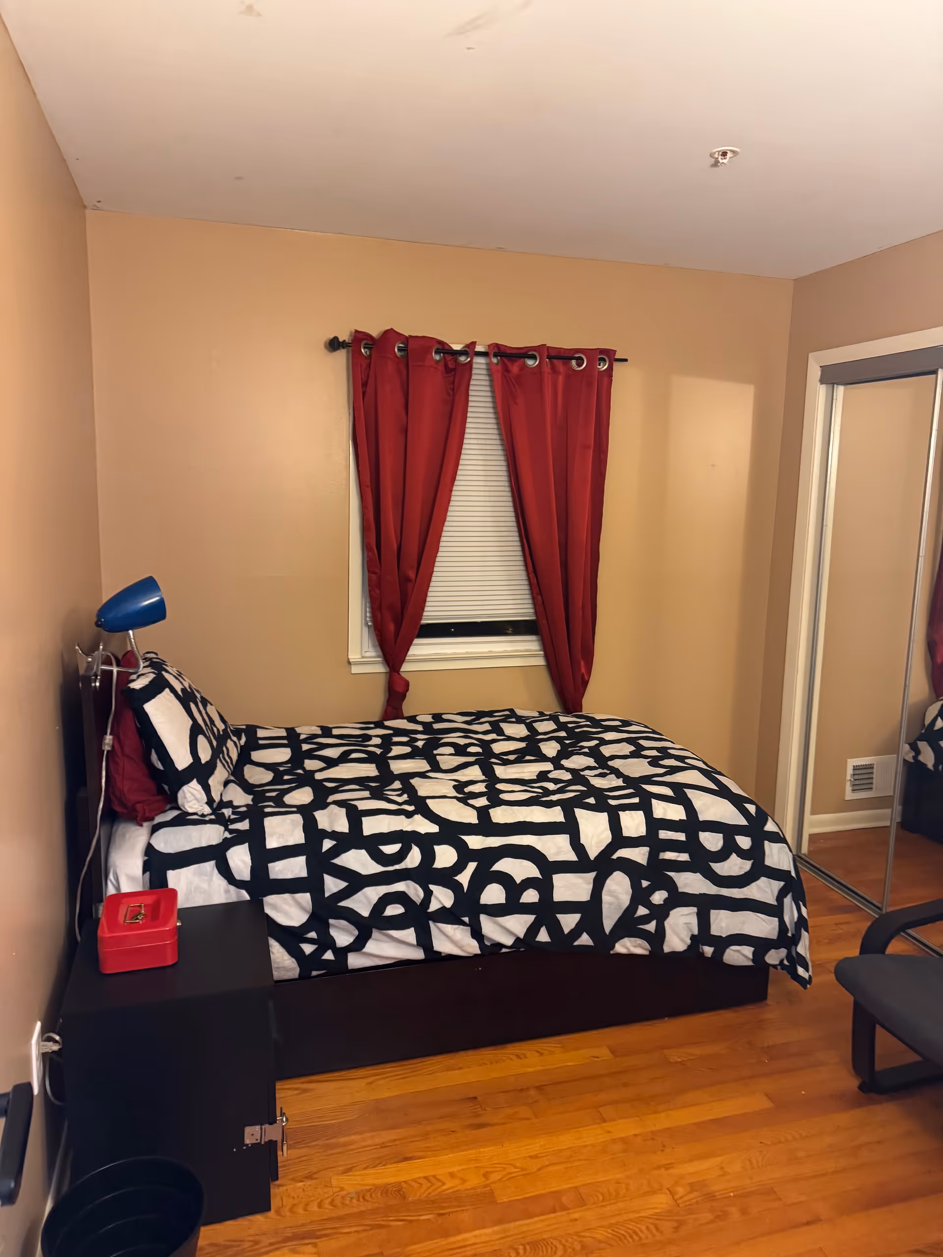 Cozy bedroom with a patterned duvet, red curtains, blue lamp, and mirrored closet.