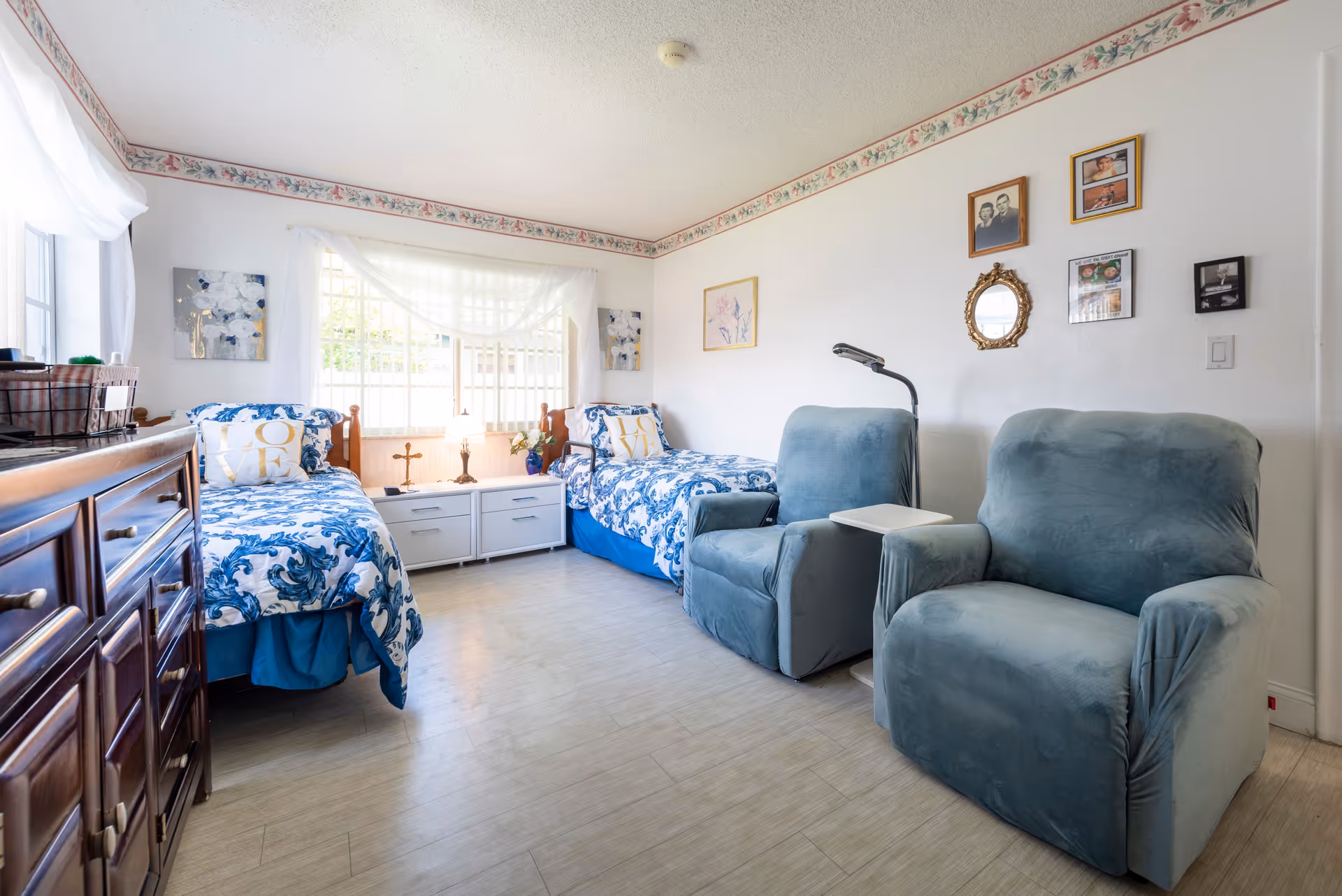 A bright and cozy bedroom in an assisted living facility featuring two twin beds with blue and white patterned bedding and decorative pillows. Between the beds is a white nightstand with a lamp and small decor items. Two comfortable blue armchairs with a small table between them are positioned against the opposite wall. The room has light-colored flooring, white walls adorned with framed pictures and a small mirror, and a large window with sheer white curtains allowing natural light to fill the space.