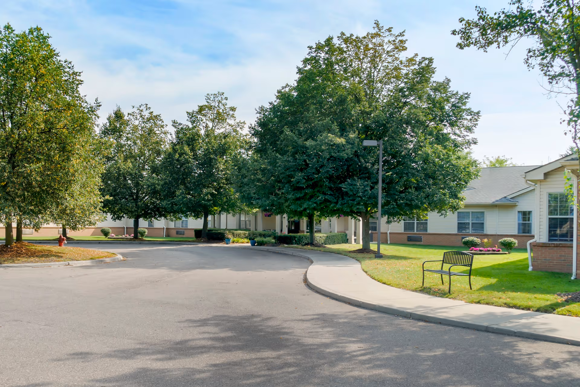 A curved driveway leading to the entrance of a single-story building surrounded by green trees and well-maintained lawns. There is a black metal bench on the grass near the sidewalk, and the sky is partly cloudy with blue patches.