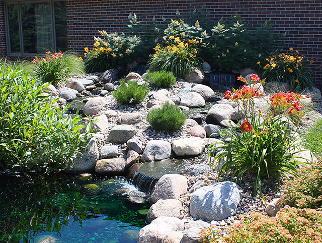 Rocky outdoor garden with a small stream, landscaping stones, and flowering plants along a brick building.