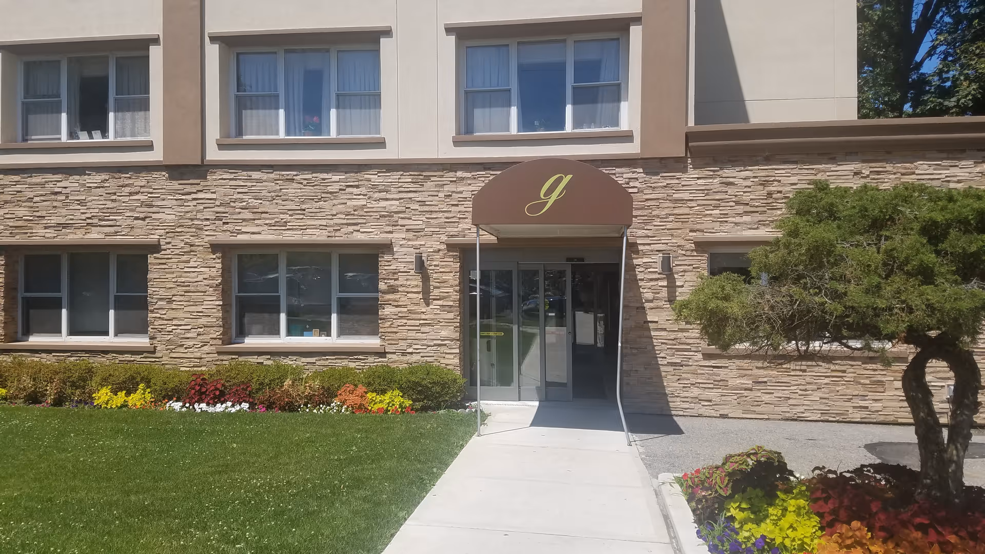 Front exterior of a senior living facility with stone facade, glass doors, and landscaped walkway.