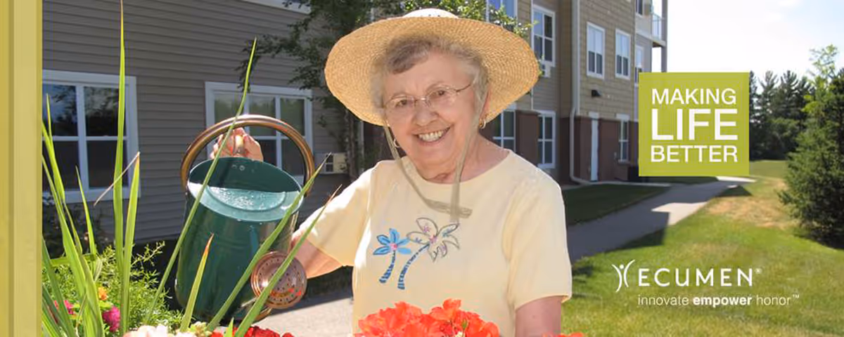 An elderly woman wearing a straw hat and glasses is smiling while watering flowers in a garden outside a senior living facility building. The building and a paved walkway are visible in the background. There is a green sign with the text 'MAKING LIFE BETTER' and the Ecumen logo with the words 'innovate empower honor' on the right side of the image.