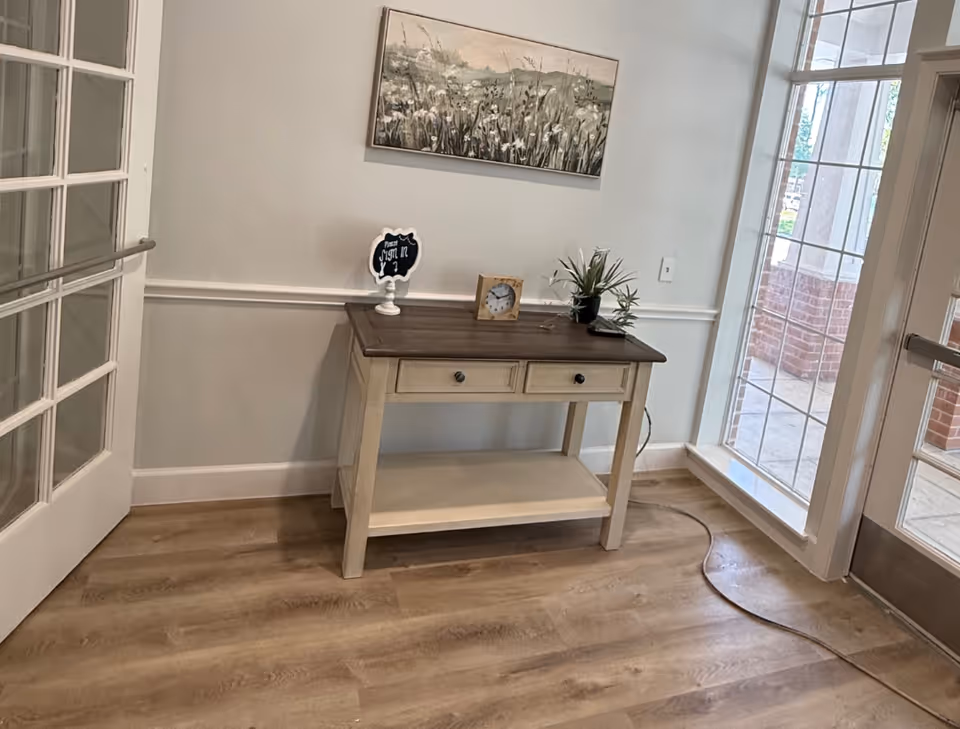 A small wooden console table with two drawers and a lower shelf is placed against a light gray wall. On top of the table are a small clock, a potted plant, and a decorative sign. Above the table hangs a landscape painting of a field with flowers. To the right, there is a large window and a glass door leading outside. To the left, a white door with glass panes is partially open. The floor is wood with a light finish.