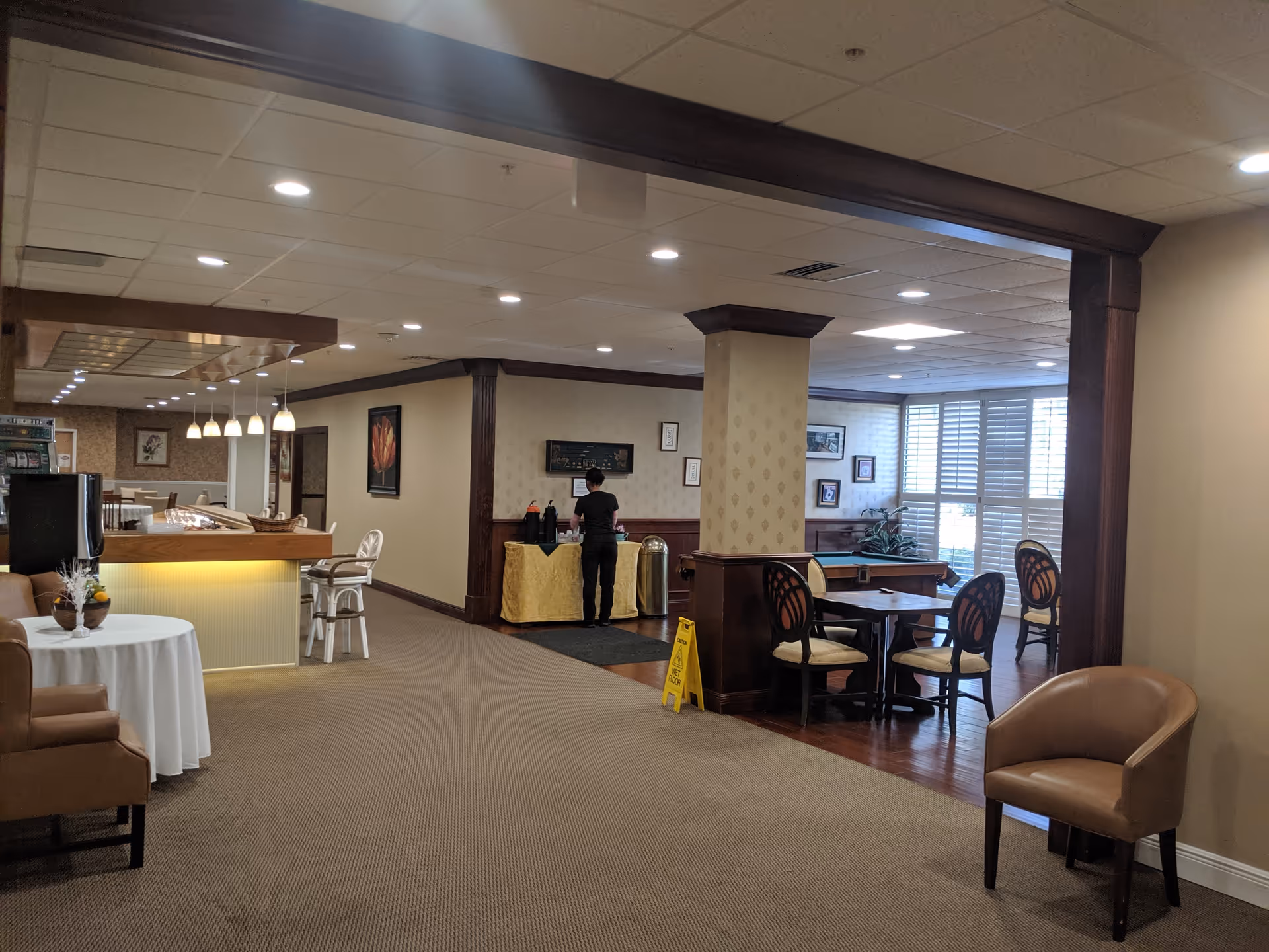 Interior common area at Plantation Oaks Senior Living with dining tables, seating, and a staff member.