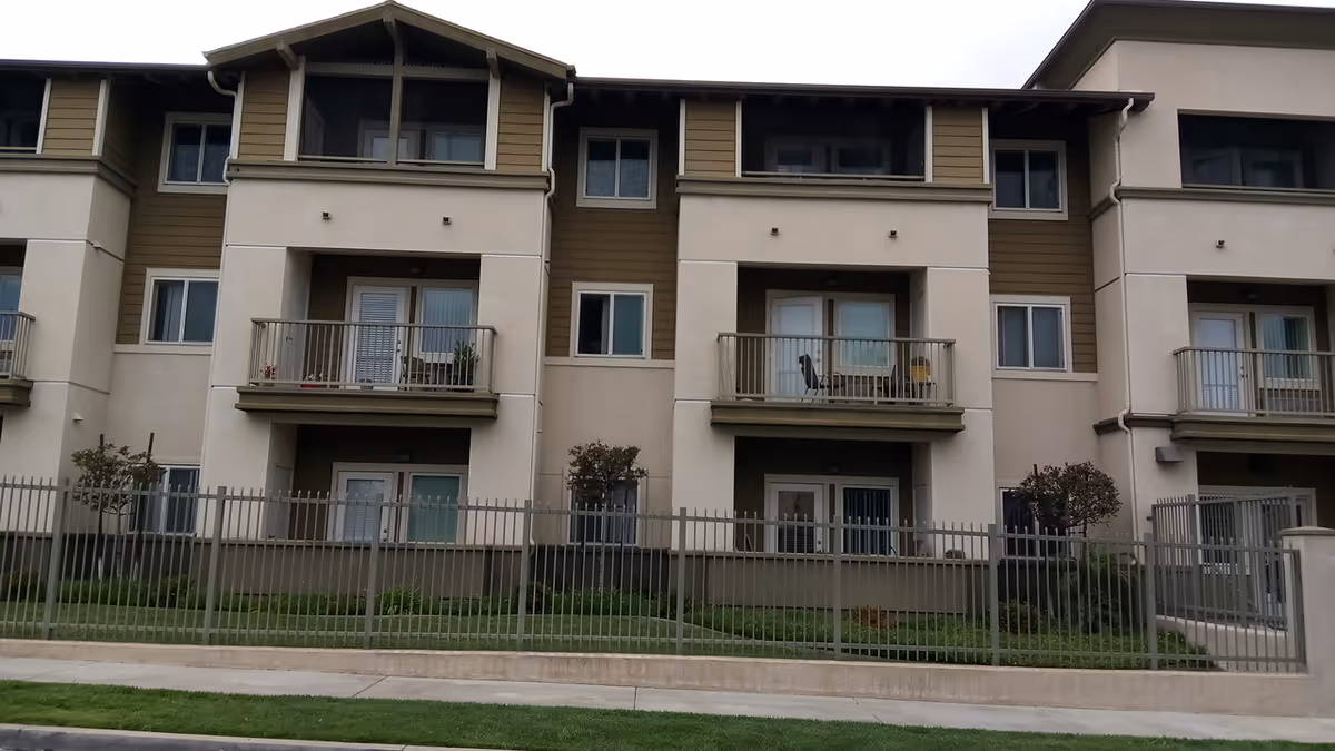 Exterior view of a multi-story senior housing building with balconies and a fenced lawn.