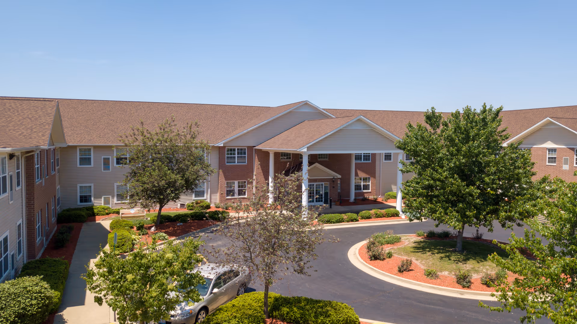 Exterior view of Bowman Estates of Danville, showing a two-story building with a covered entrance, surrounded by trees, shrubs, and a circular driveway with a parked car under a clear blue sky.