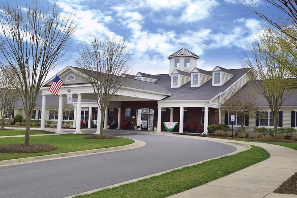 Front exterior view of a senior living facility building with a covered entrance supported by white columns, an American flag, and a circular driveway. The building has a combination of brick and siding with multiple windows and a cupola on the roof. Leafless trees and green grass surround the driveway and sidewalk.