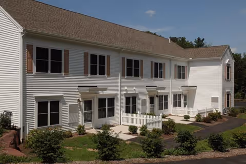 Exterior view of a two-story white residential building with multiple windows and small fenced patios. The building is surrounded by landscaped greenery and a paved walkway under a clear blue sky.