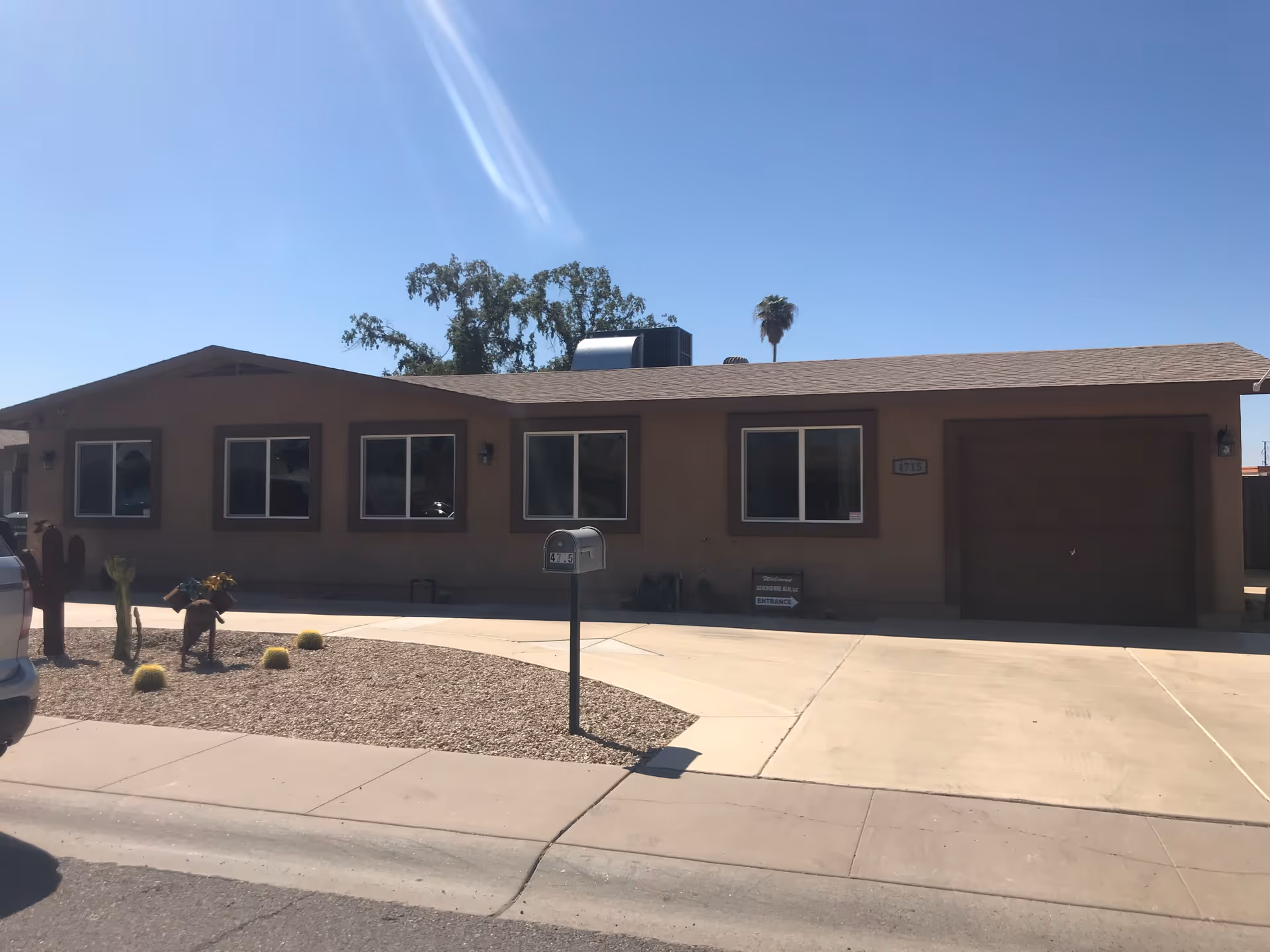 Single-story building with a brown exterior, four windows, a garage door, and a mailbox in front. The area in front of the building has a gravel yard with some small desert plants and a concrete driveway under a clear blue sky.