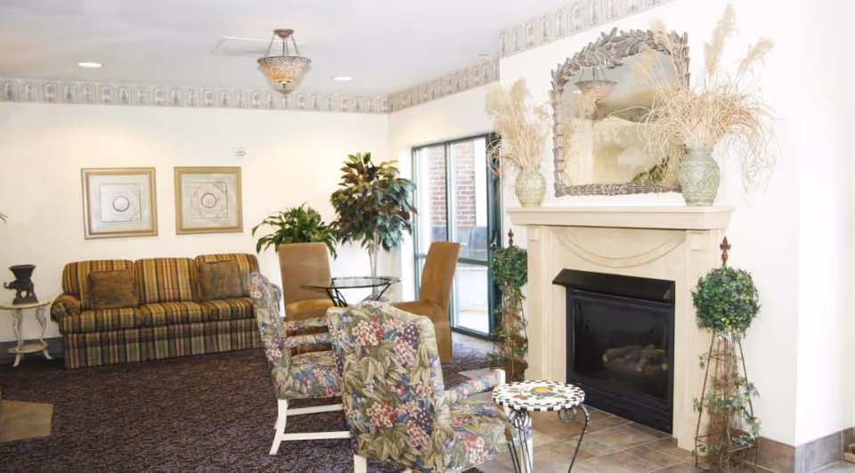 A bright common room with a fireplace, mirror, floral armchairs, a striped sofa, decorative plants, and a small table.