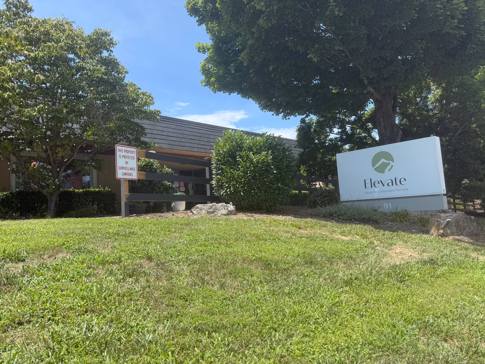 Exterior view of a senior care facility with sign, shrubs, and trees.