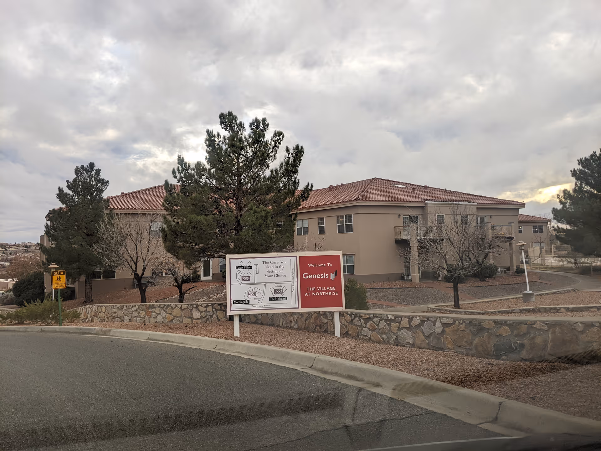 Exterior view of a beige, tile-roofed senior living building with a stone wall and sign.