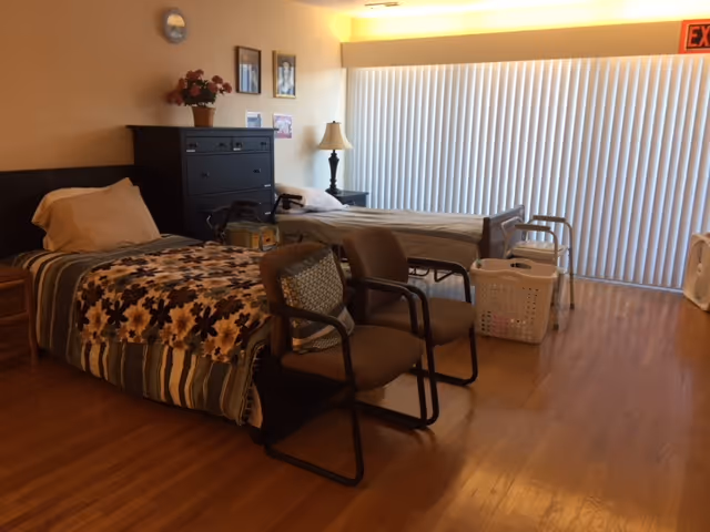 A room with two single beds, each with pillows and blankets. There are two chairs with cushions placed between the beds. A black dresser with a flower pot on top is against the wall, along with a lamp on a small table. Vertical blinds cover a large window or sliding door, and a walker and laundry basket are near one of the beds. The floor is wooden, and an exit sign is visible above the blinds.