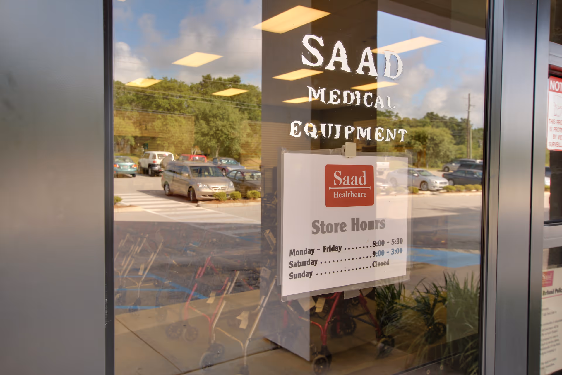 Glass storefront displaying Saad Medical Equipment sign with store hours, reflecting parked cars.