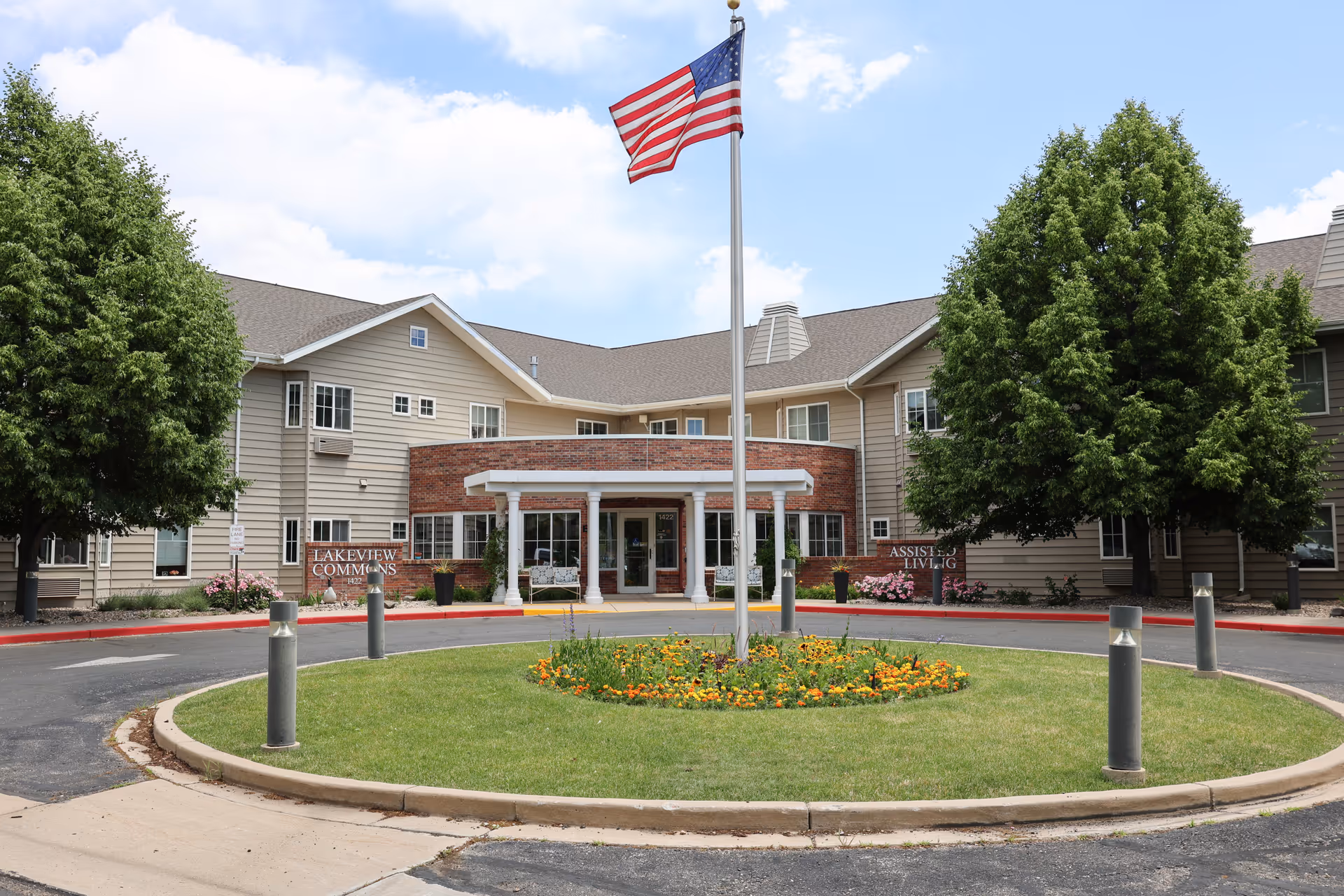 Front exterior view of Lakeview Commons assisted living facility with a circular driveway, a flagpole with an American flag in the center, and trees and flowers surrounding the entrance.