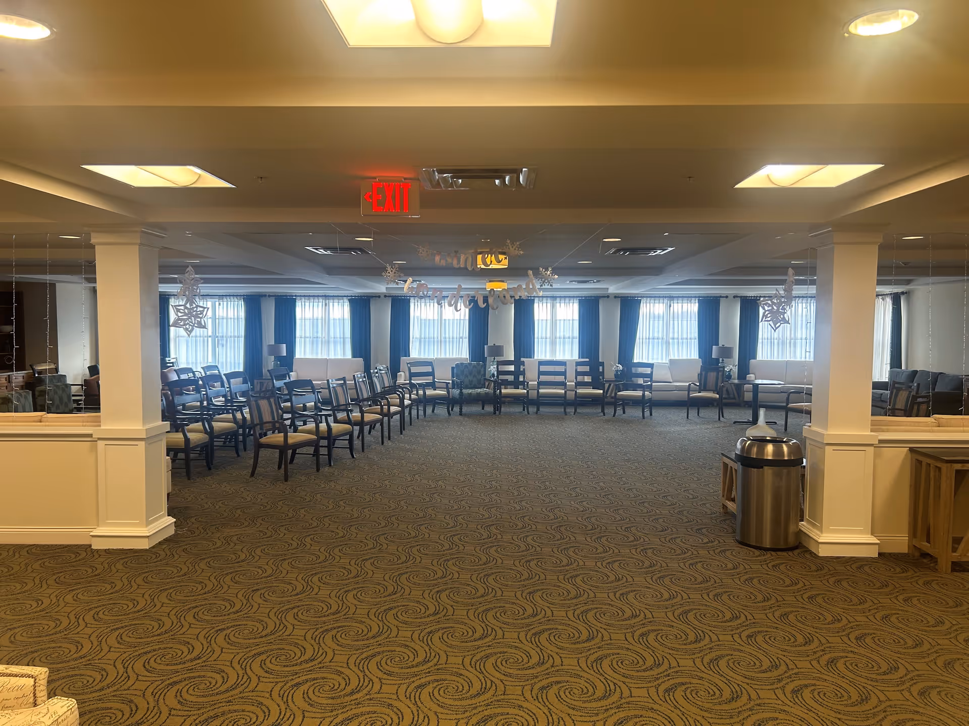 A large, carpeted common area in a senior living facility with rows of chairs arranged in a semi-circle facing the center. The room has large windows with blue curtains, white sofas, and several lamps. There are decorative snowflakes hanging from the ceiling and a banner that reads 'winter wonderland'. The ceiling has recessed lighting and an illuminated exit sign is visible above the chairs.