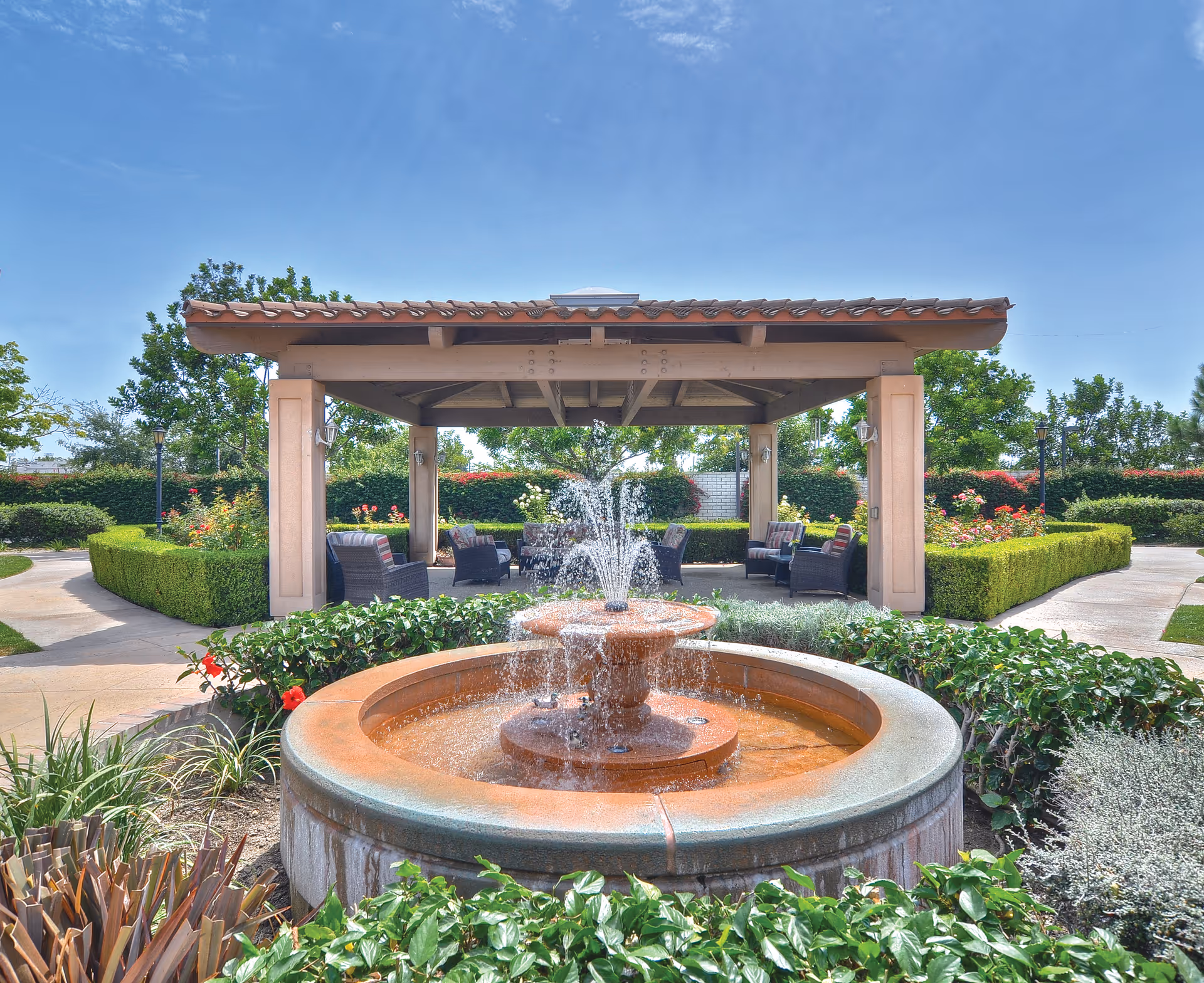 Outdoor garden area with a circular water fountain in the foreground and a covered seating area with cushioned chairs under a wooden pergola. The garden is surrounded by neatly trimmed hedges, flowering plants, and trees under a clear blue sky.