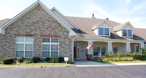 Front exterior of a single-story brick-and-siding senior living building with a covered porch, American flag, and landscaping.
