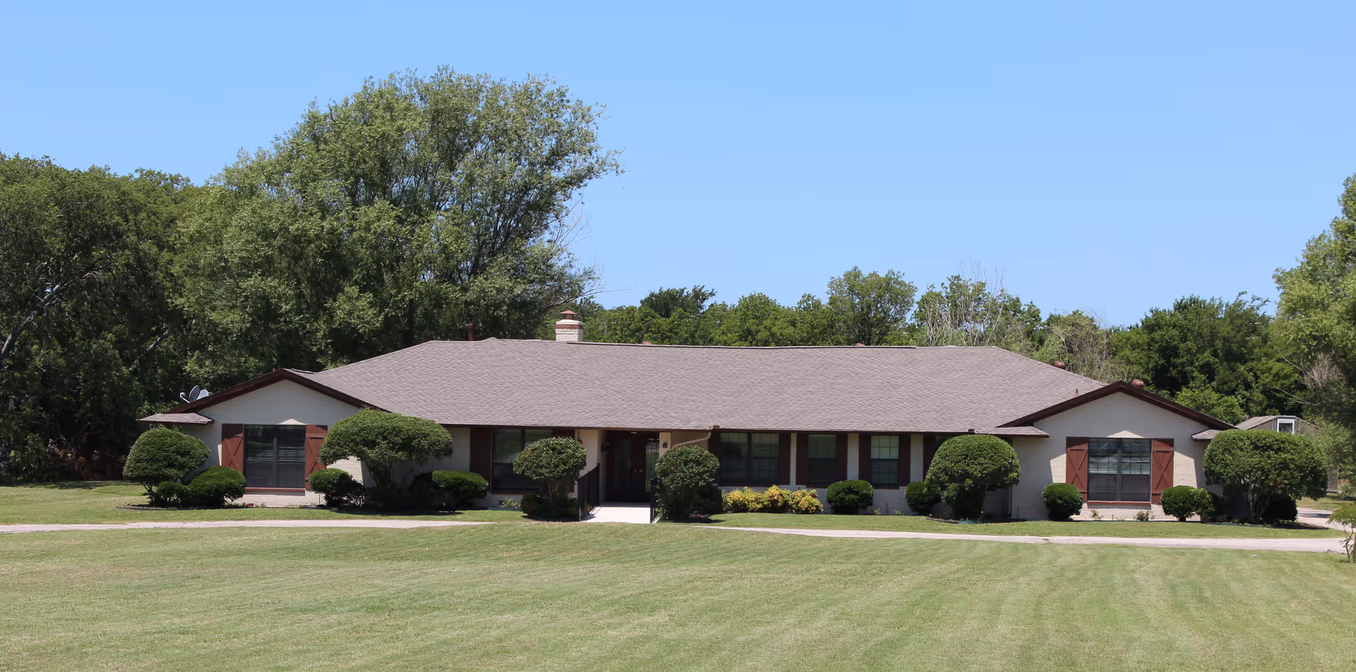 Single-story building with a brown roof and beige walls surrounded by neatly trimmed bushes and trees under a clear blue sky, with a large green lawn in the foreground.