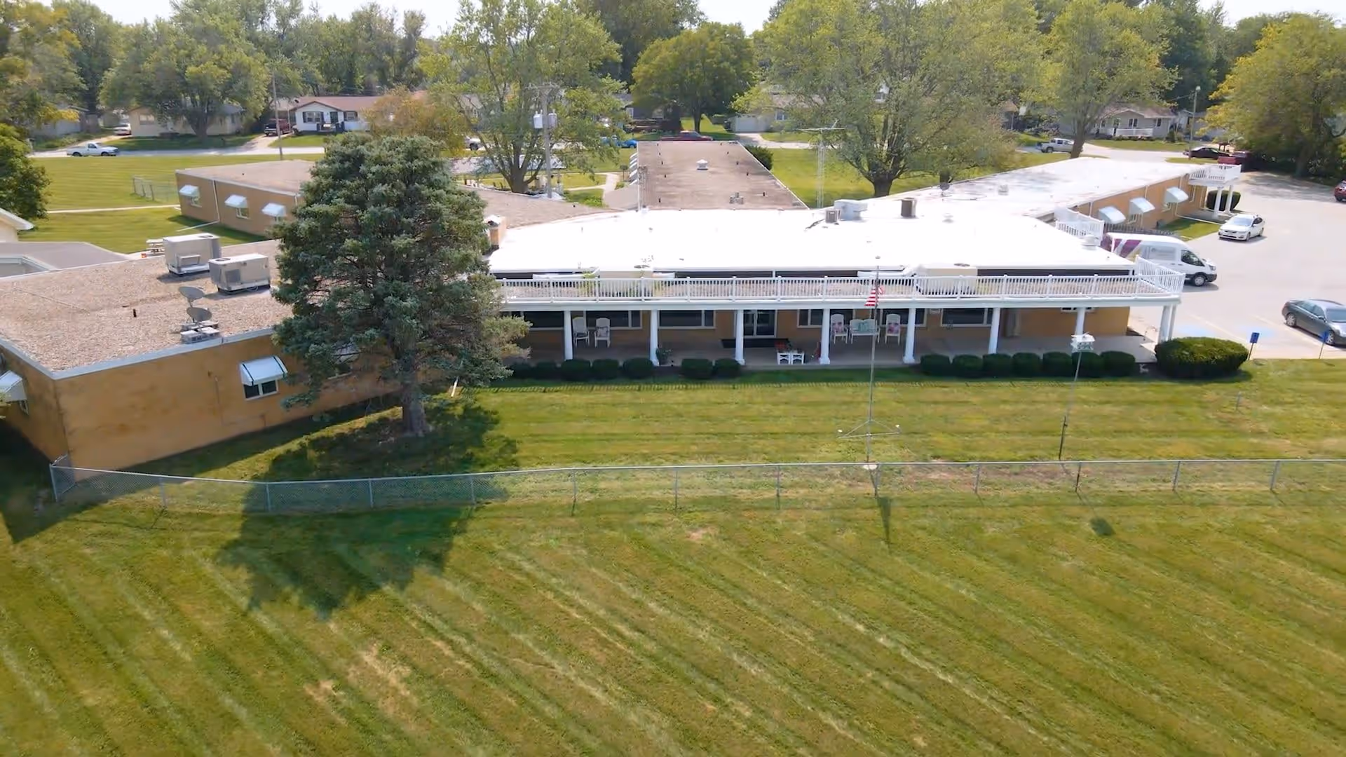 Aerial view of a single-story building with a covered front porch, fenced grassy lawn, and nearby parking.