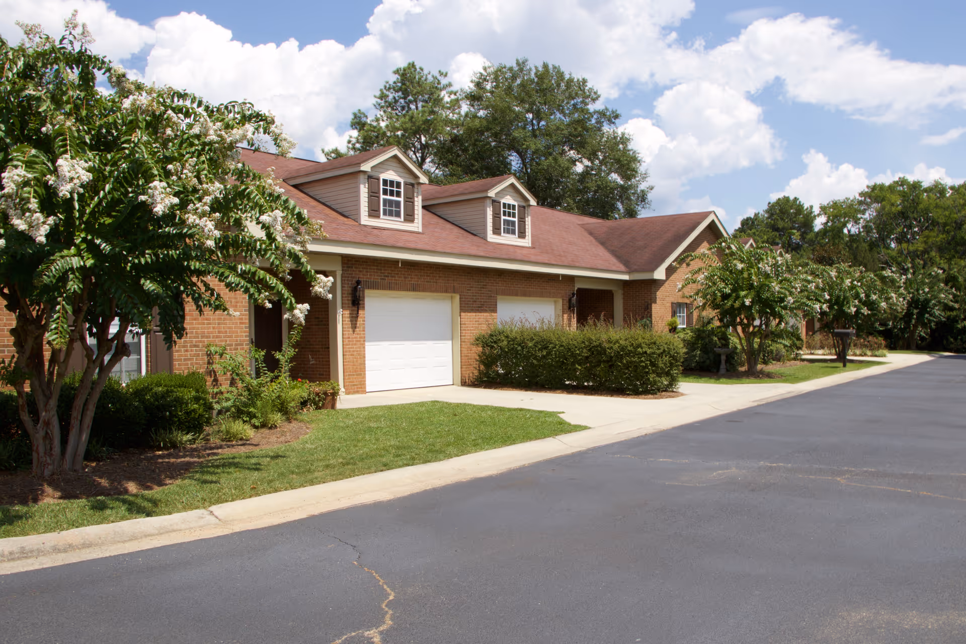 Exterior view of a single-story brick building with a red roof and two white garage doors, surrounded by green bushes and trees under a partly cloudy sky.