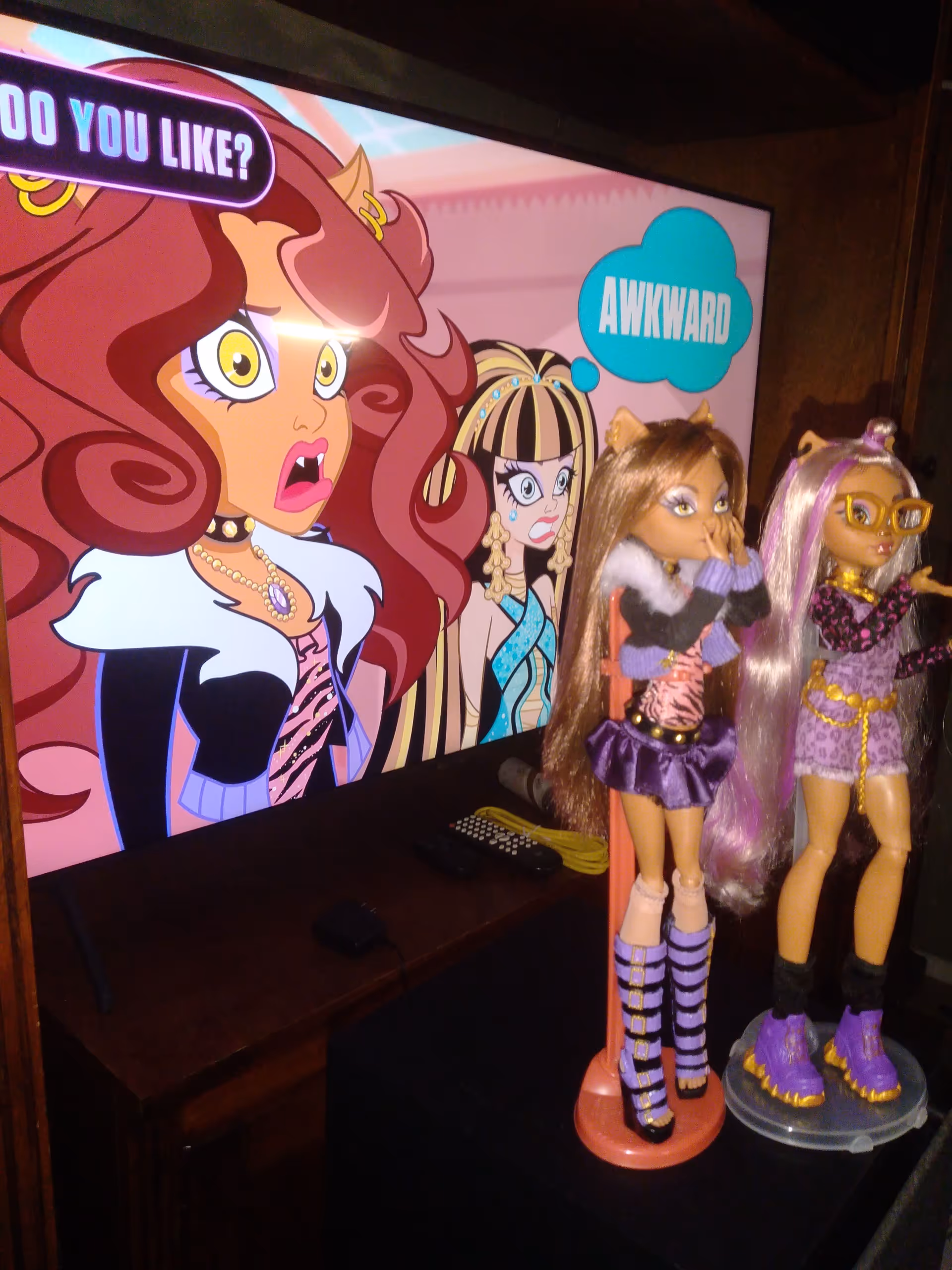 TV displays a colorful cartoon; two fashion dolls stand beside a dark table.