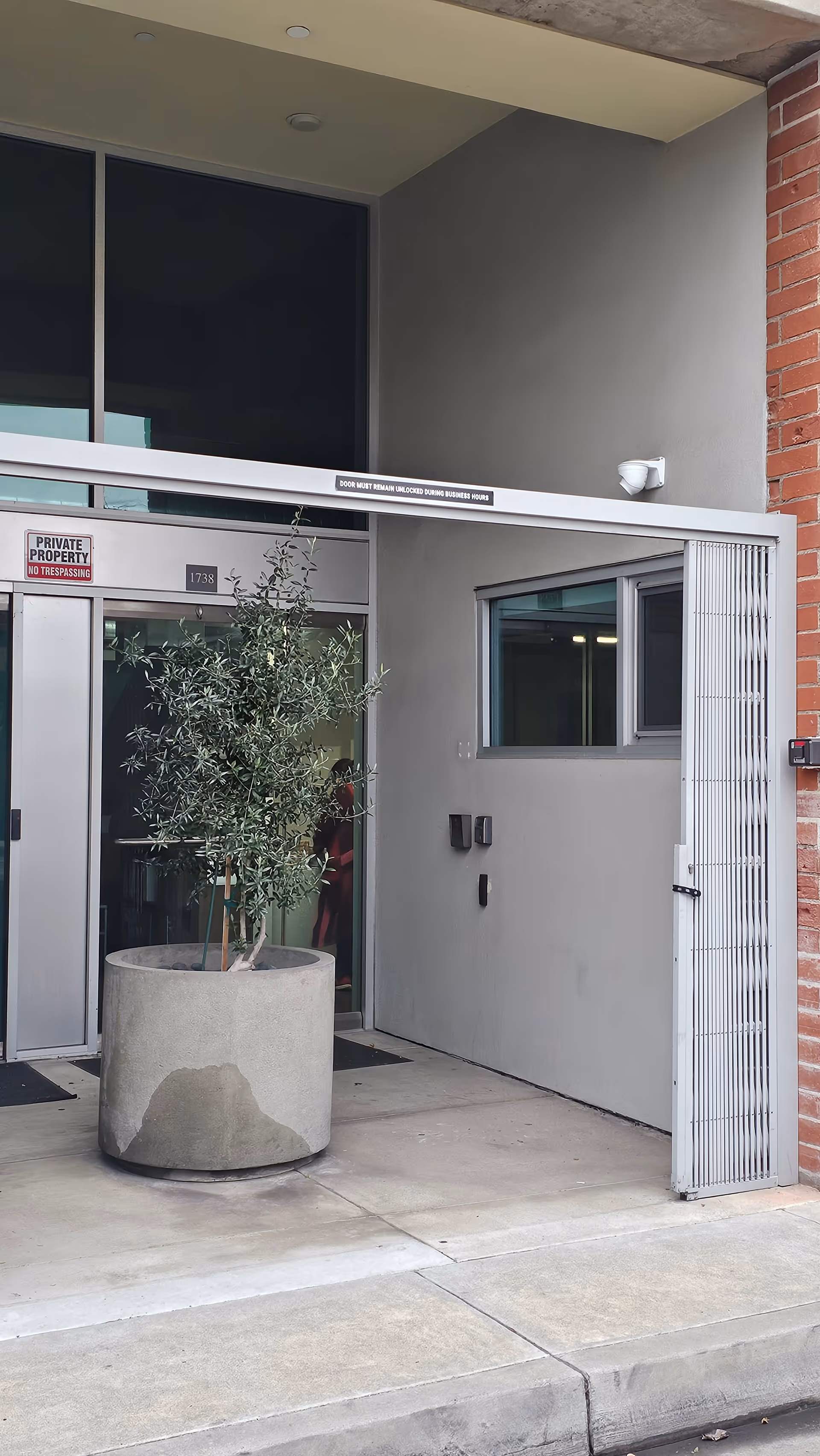 Exterior entrance to Tandem Care facility with glass doors, a large planter olive tree, and sign.