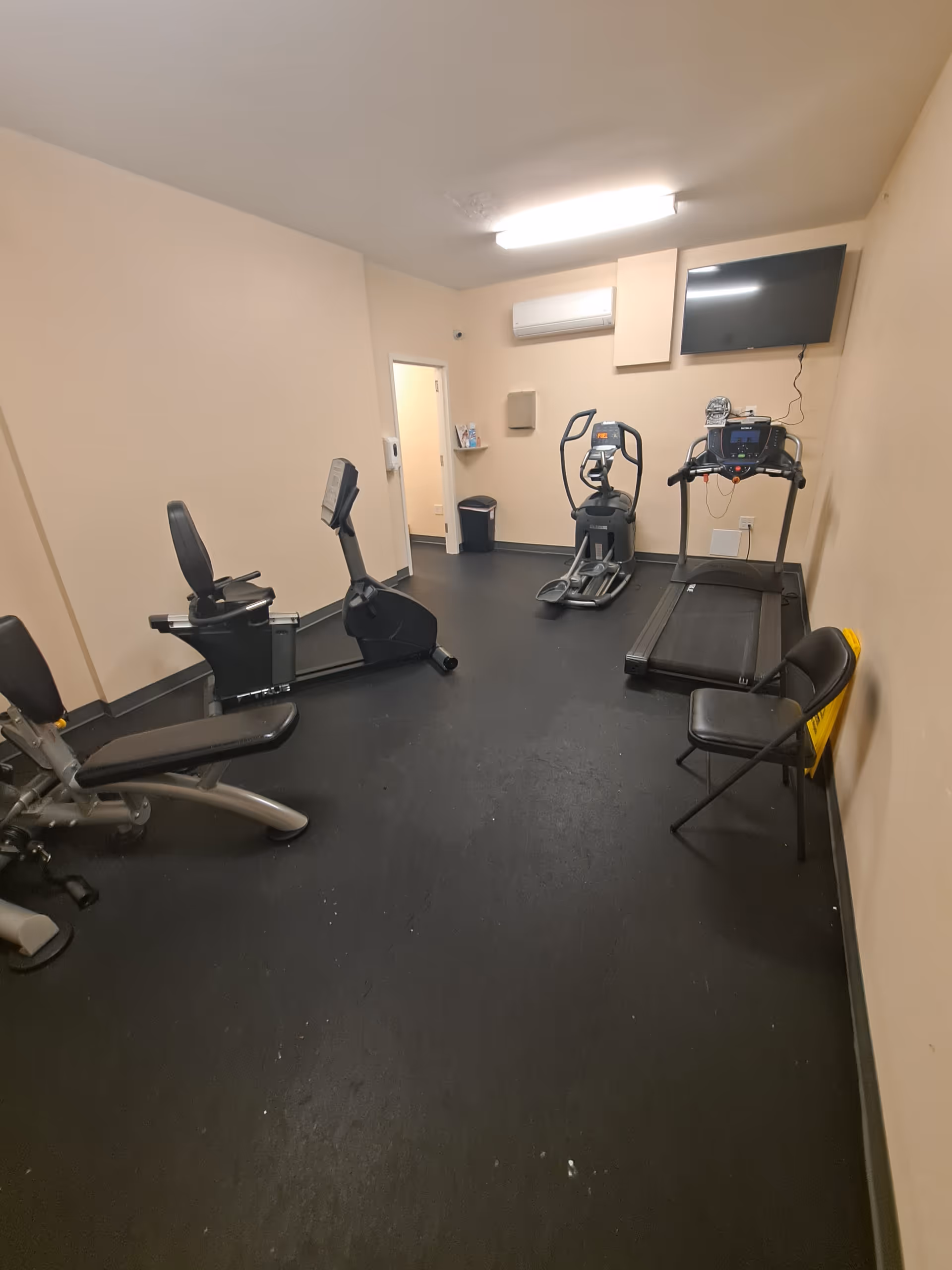 Small gym room with cardio machines, a wall-mounted TV, and beige walls.