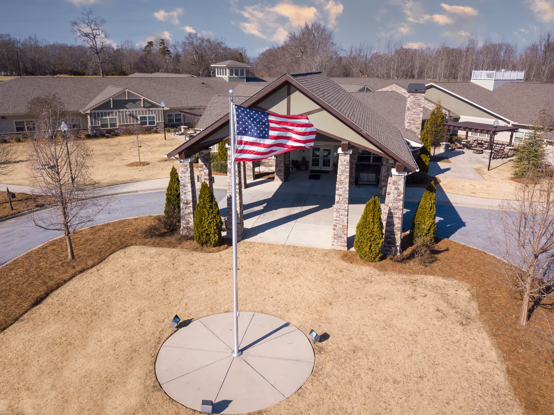 Front exterior view of a senior living facility with a covered entrance supported by stone pillars, an American flag on a flagpole in the foreground, and surrounding dry grass and trees.
