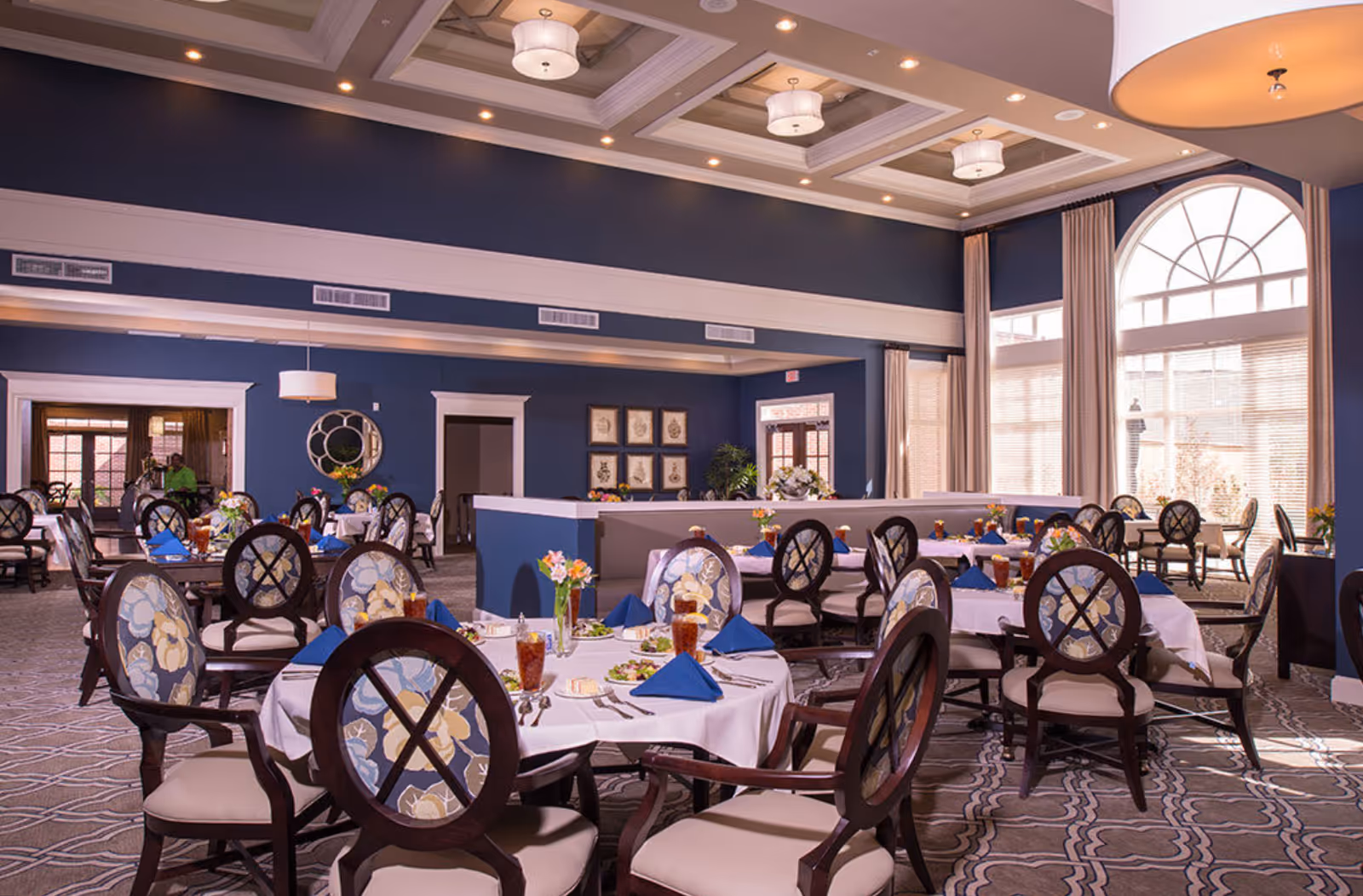 A spacious dining room with multiple round tables covered in white tablecloths, each set with blue napkins, glasses of iced tea, plates of salad, and silverware. The chairs have dark wooden frames with floral-patterned upholstery. The room features high ceilings with recessed lighting and large arched windows letting in natural light. The walls are painted dark blue with white trim, and there are decorative elements such as framed artwork and a round mirror.