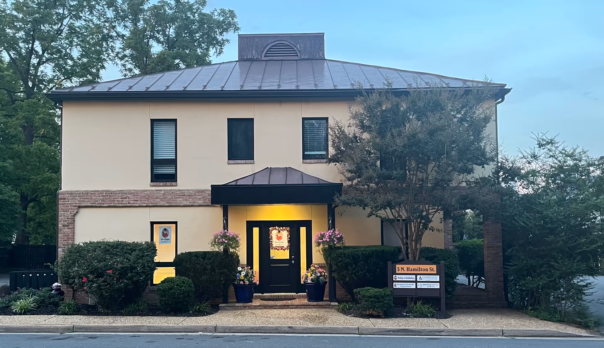 Two-story beige building with brick accents, black doorway canopy, and potted flowers.