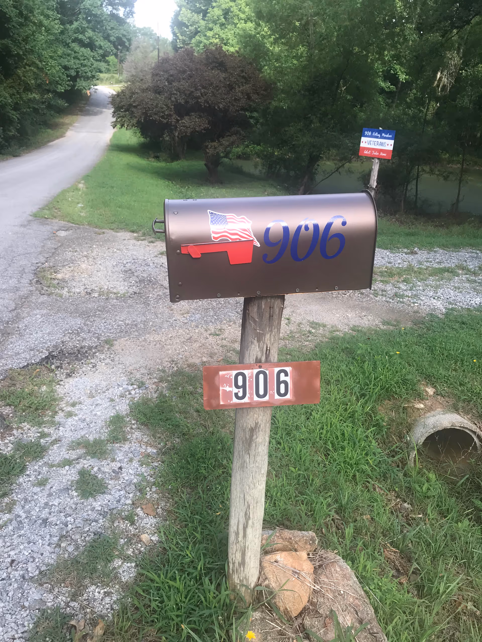 Brown mailbox with 906 address on a gravel driveway amid trees and a Veterans sign.