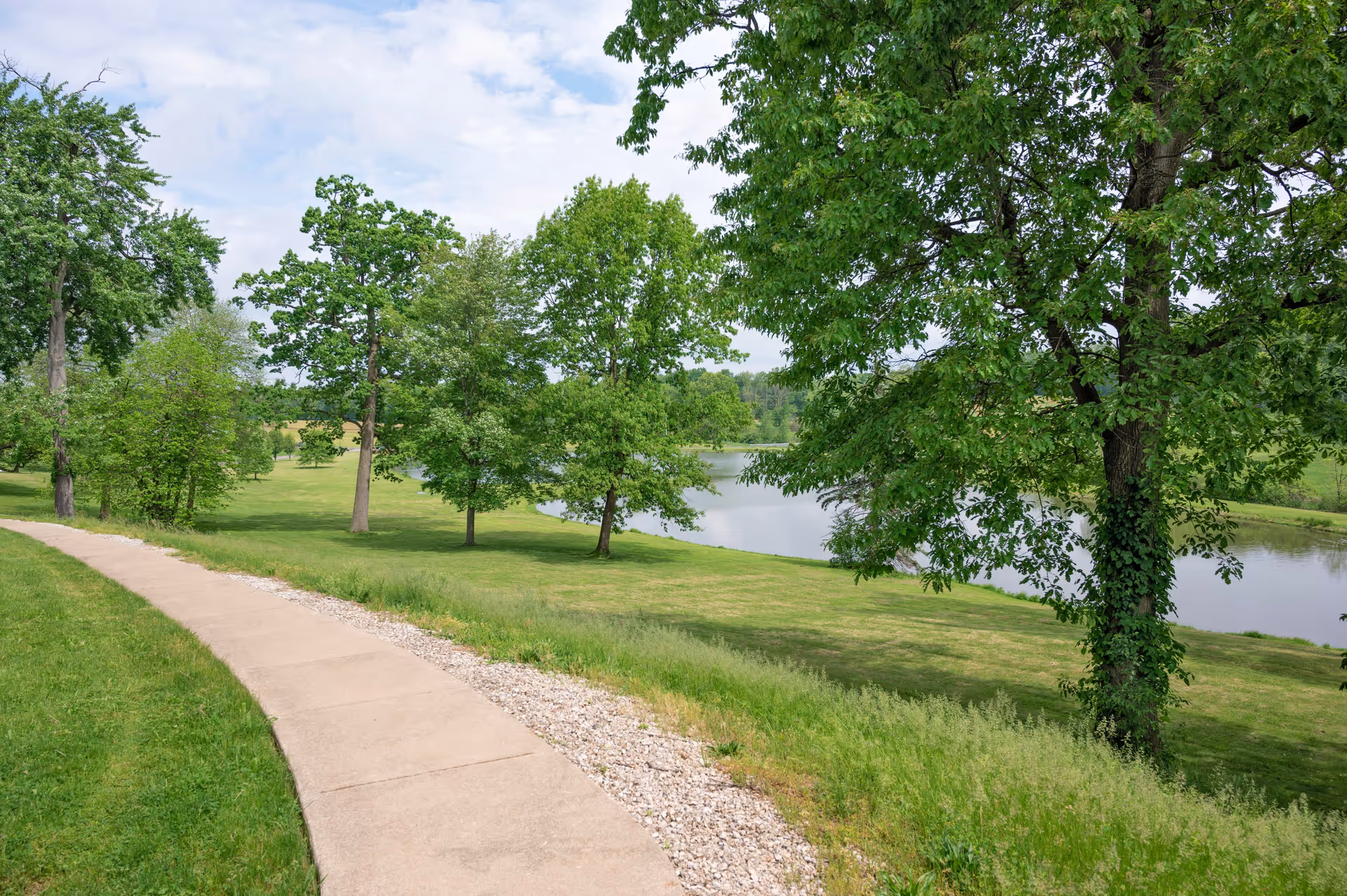 A paved walking path curves through a green grassy area with several trees and a small body of water visible in the background under a partly cloudy sky.