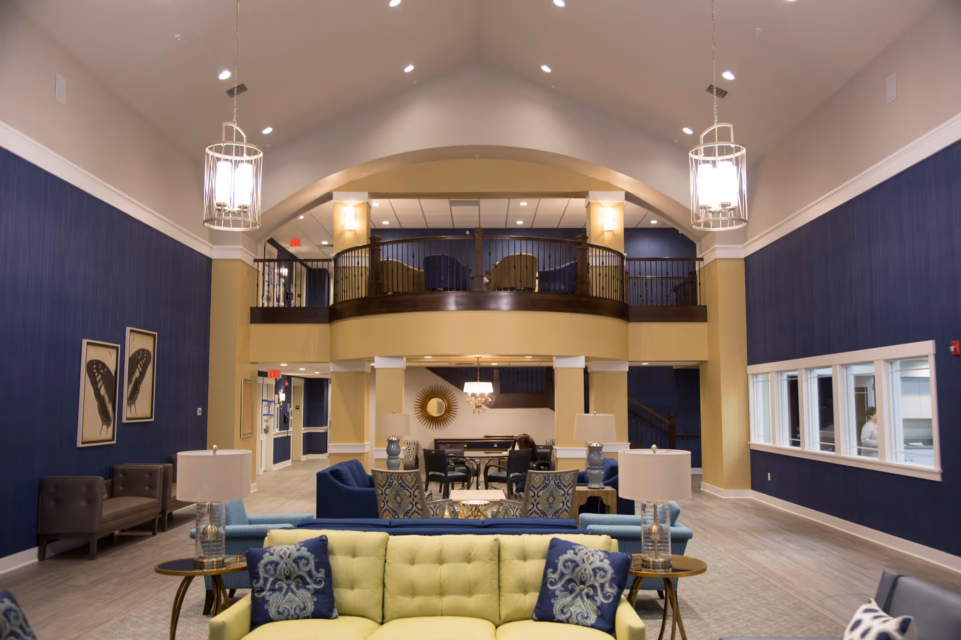 Spacious senior living facility common area with high vaulted ceilings, blue and beige walls, multiple seating arrangements including sofas and armchairs, decorative lamps, and a balcony with additional seating above.
