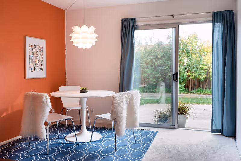 A bright dining area with round white table, blue rug, fur-covered chairs, and sliding glass door.