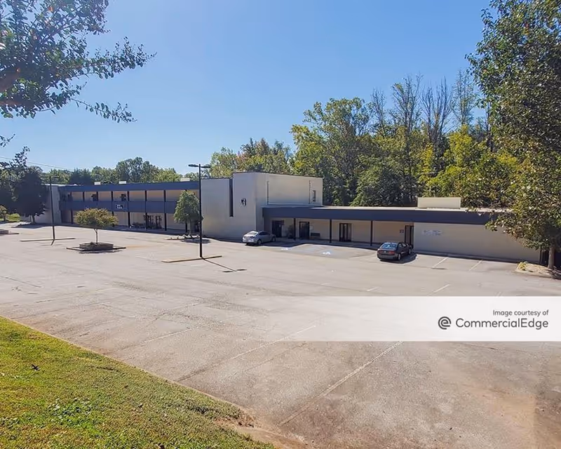 Exterior view of a single-story commercial building with a large parking lot and trees.