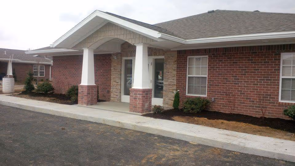 Front exterior of a single-story brick building with a covered columned entrance, windows, sidewalk, and small landscaped beds.