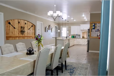 Open dining area with a long table and upholstered chairs, a chandelier overhead, and a bright white kitchen visible in the background.