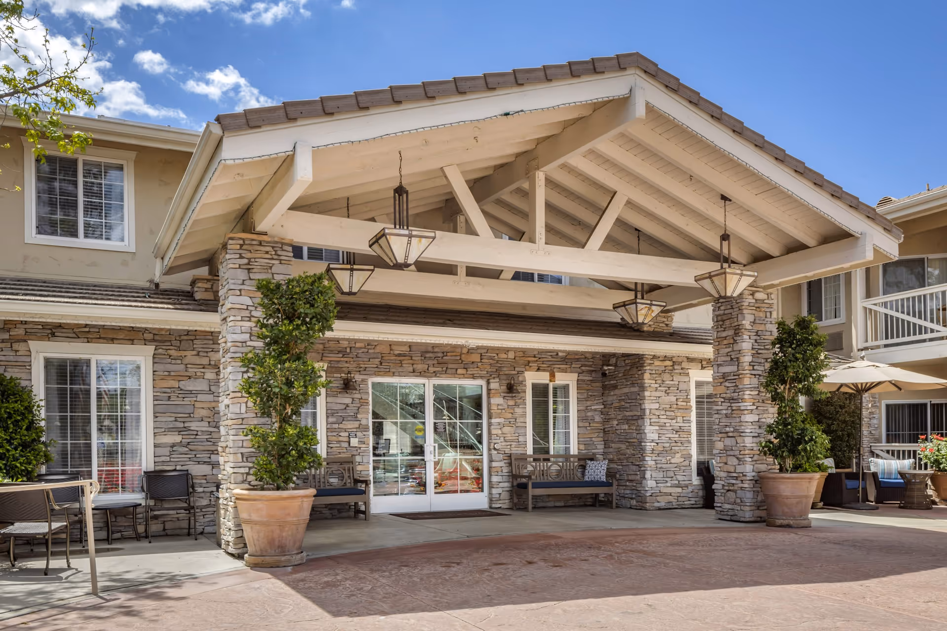 Covered stone-and-timber entrance with hanging lanterns, potted trees, benches and double glass doors under a pitched roof.
