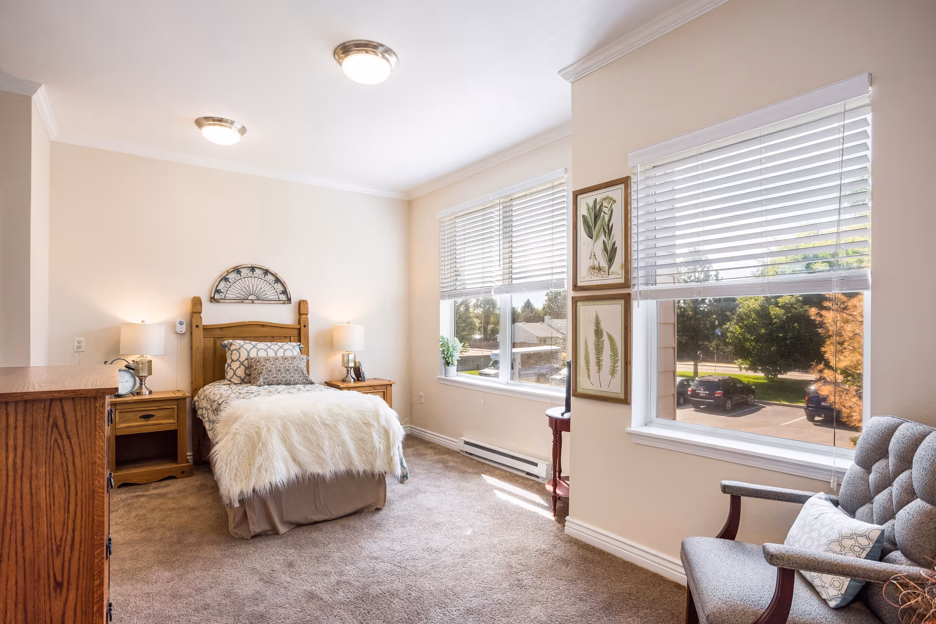 A bright bedroom with a single wooden bed covered with a white fluffy blanket and decorative pillows. There are two wooden nightstands with lamps on either side of the bed. A wooden dresser is on the left side of the room. Two large windows with white blinds let in natural light, showing a view of parked cars and trees outside. A gray upholstered armchair with a patterned pillow is positioned near the windows. The walls are light-colored, and two framed botanical prints hang between the windows.
