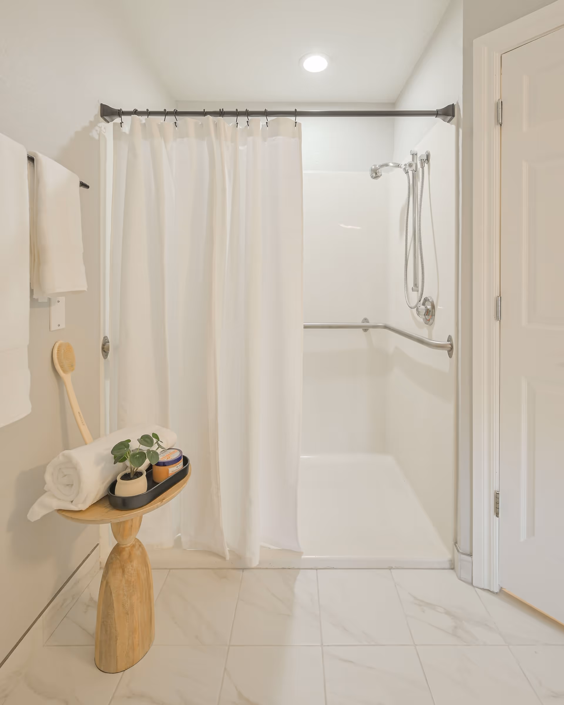 A clean and bright bathroom featuring a walk-in shower with a white curtain, a silver showerhead, and a grab bar. Next to the shower is a wooden stool holding a rolled white towel, a small potted plant, and some bath products. White towels hang on a towel rack on the wall. The floor is tiled with light-colored tiles.