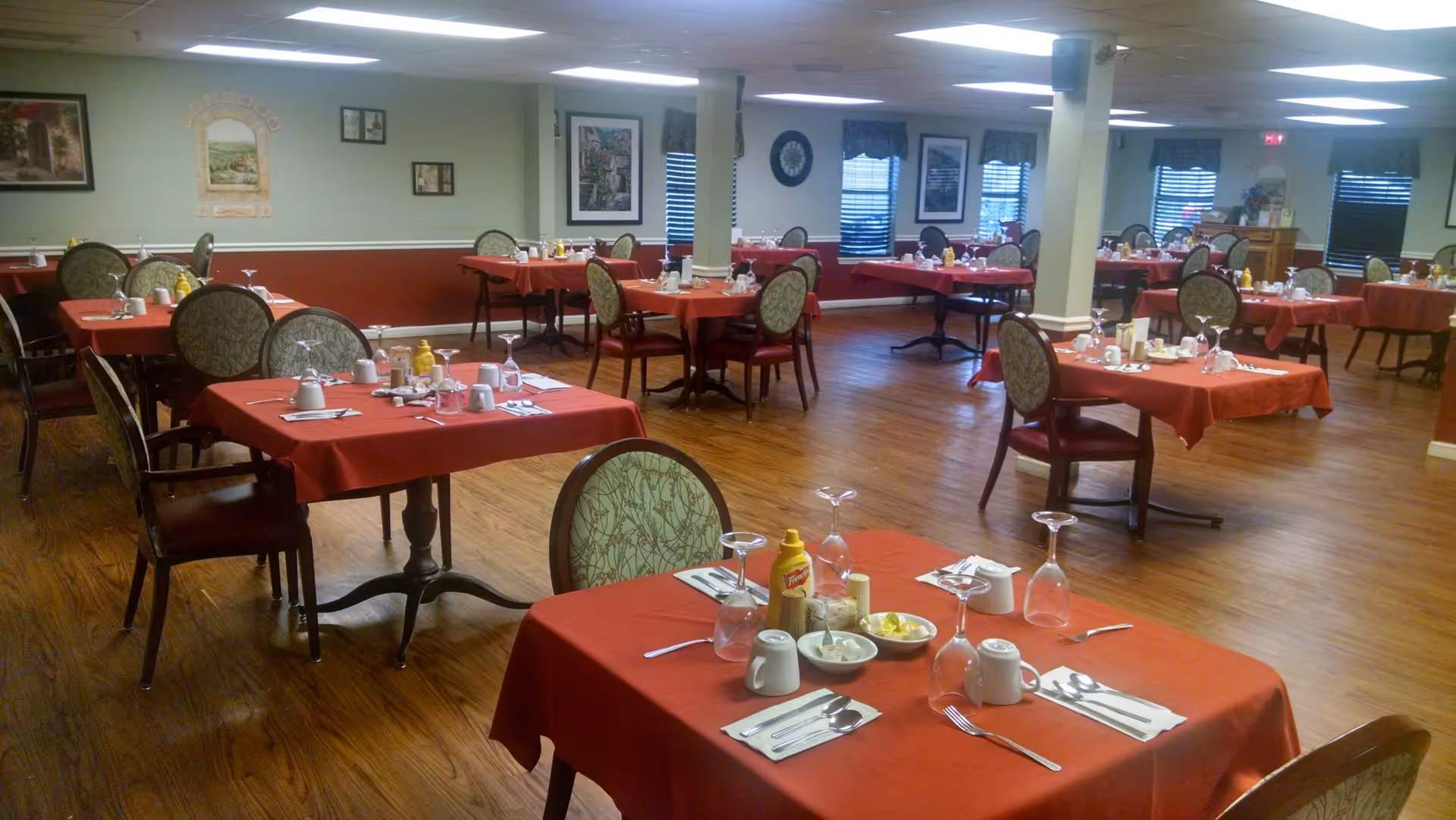Spacious dining room with multiple tables covered in red tablecloths set with utensils and condiments.