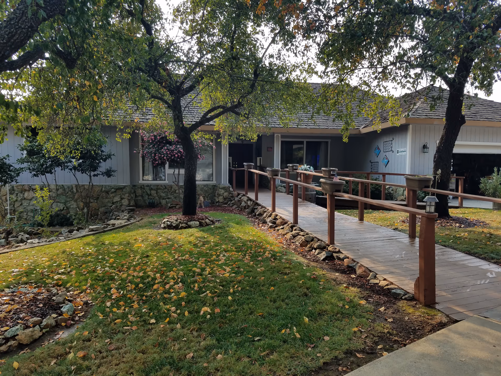 Wooden ramp walkway to Oak Grove Senior Care, with trees, lawn, and a low stone border.