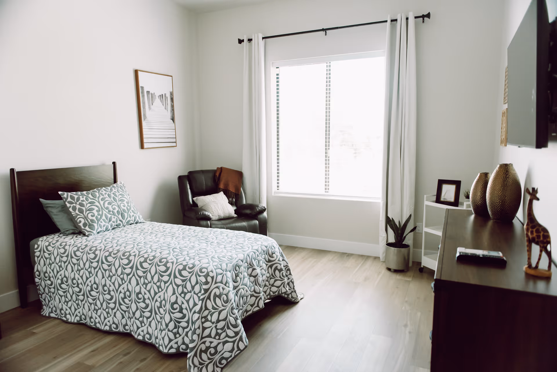Bright, minimally furnished bedroom with a patterned single bed, armchair, window with curtains, and a dresser with decor.