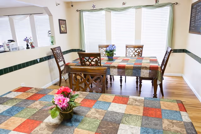 Bright communal dining room with two tables covered in colorful patchwork tablecloths, wooden chairs, floral centerpieces, and large sunlit windows.