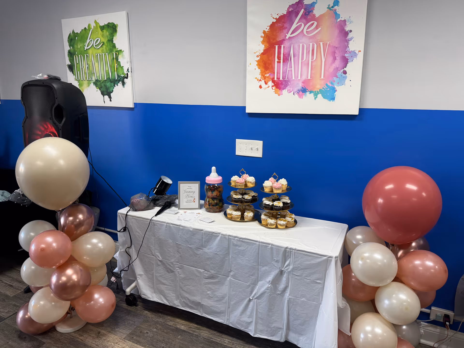 A dessert table with cupcakes, a pink bottle, and balloons against a blue wall.