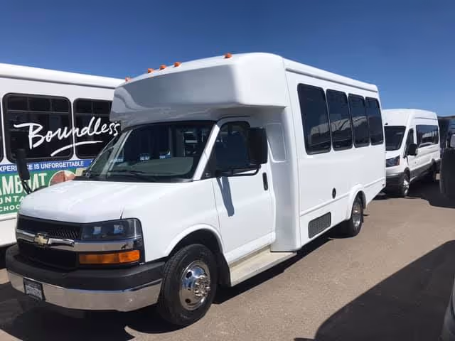 A white shuttle bus parked outdoors on a sunny day with other similar vehicles in the background.